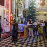 Na zdjęciu jest grupa muzyków w tradycyjnych strojach w kościele. Obok stoi udekorowana choinka. W tle znajdują się złote detale wnętrza.