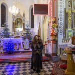 Kościół z trzema osobami na scenie. Jasny ołtarz w tle, choinki z lampkami. Mężczyzna z mikrofonem, kobieta i muzyk z skrzypcami.