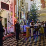 Wnętrze kościoła z choinką i kolędnikami w strojach ludowych. Ściany zdobią freski i złote elementy. Przy mikrofonie stoi mężczyzna w ciemnym ubraniu.