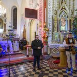 Grupa mężczyzn w kościele na tle ołtarza i choinek. Ornaments stylu gotyckiego i jasne światła. Na podłodze czarno-biała szachownica.