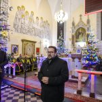 Mężczyzna w czarnym płaszczu przemawia przy mikrofonie w kościele. Tło zdobią choinki z kolorowymi lampkami i freski. Wnętrze ma jasne ściany i dekorowane ołtarze.