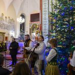 Ludzie w strojach ludowych grają na instrumentach w kościele. W tle choinki z kolorowymi światełkami. Atmosfera świąteczna.