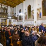 Na zdjęciu widać wnętrze kościoła z dużą liczbą osób siedzących na ławkach. Ściany są ozdobione malowidłami i napisami. Wiszące żyrandole oświetlają przestrzeń ciepłym światłem.