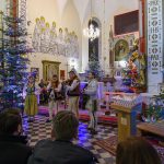 Grupa muzyków w strojach ludowych gra w kościele, otoczona ozdobnymi choinkami. Ściany zdobią freski, a wnętrze jest bogato oświetlone. Widownia siedzi na czarno-białej podłodze.