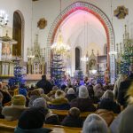 Kościół wypełniony ludźmi, udekorowany świątecznymi choinkami. W tle ołtarz, freski i żyrandole. Dominują kolory bieli, złota i zieleni.