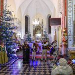 Grupa muzyków w ludowych strojach gra w kościele. W tle choinki, dekoracje i złote detale na ścianach. Publiczność siedzi i robi zdjęcia.