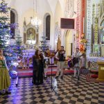 W kościele ozdobionym choinkami z kolorowymi światełkami występuje zespół muzyczny w tradycyjnych strojach. Tło jest barwne, z detalami architektonicznymi i religijnymi. Kobieta w zielonej sukni stoi z boku