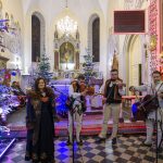 Cztery osoby w tradycyjnych strojach góralskich grają na instrumentach w udekorowanym kościele. Zieleń choinek z kolorowymi lampkami i złote detale ołtarza tworzą świąteczny nastrój. W tle widać białe ściany