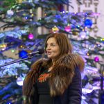 Kobieta w futrzanym płaszczu mówi do mikrofonu. W tle widoczna choinka z kolorowymi lampkami i bombkami. Świąteczna atmosfera.