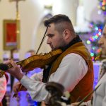 Mężczyźni grają na skrzypcach w tradycyjnych strojach. Tło zdobią świąteczne światła. Scena rozgrywa się w eleganckim wnętrzu.