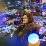 Na zdjęciu widoczna jest kobieta w futrzanej pelerynie przy mikrofonie. W tle ozdobiona choinka z kolorowymi bombkami i światełkami. Atmosfera świąteczna z ciepłymi, świątecznymi kolorami.