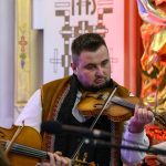 Muzyk w tradycyjnej kamizelce gra na skrzypcach. Tło kolorowe z elementami złota i czerwieni. Widoczny mikrofon z przodu.