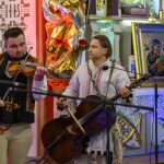 Dwóch mężczyzn gra na instrumentach smyczkowych w kolorowym wnętrzu kościoła. Tło ozdobione złotą rzeźbą i ikoną Matki Boskiej. Oświetlenie w ciepłych tonacjach.