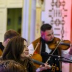 Kobieta śpiewa przy mikrofonie w tradycyjnym stroju. Za nią muzyk gra na skrzypcach. Tło w jasnych kolorach i wzorach.