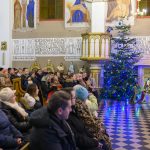 Wnętrze kościoła z siedzącymi ludźmi, ozdobiona choinka po prawej. Na ścianach religijne malowidła i teksty. Dziecko i kobieta w ludowym stroju stoją obok szopki.