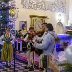 Grupa ludzi w tradycyjnych strojach góralskich gra muzykę w kościele ozdobionym choinkami. W tle ściany z freskami. Kolory: zieleń, biel, brąz.