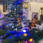 Na zdjęciu widać świąteczny koncert w kościele. Przy choince w ludowych strojach śpiewa kobieta i grają muzycy. Tło zdobią kolorowe ozdoby i religijne obrazy.