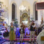 Grupa muzyków w tradycyjnych strojach występuje w udekorowanym świątecznie kościele. Ściany zdobią freski i witraże, a wnętrze oświetla żyrandol. Choinki z kolorowymi lampkami tworzą świąteczny nastrój