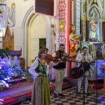 Grupa muzyków gra przed ołtarzem w kościele, ubranych w tradycyjne stroje. Obok dekoracje świąteczne z choinkami i lampkami. W tle złote ornamenty i ikony.