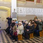 Grupa ludzi siedzi w drewnianych ławkach kościoła, obok choinka z ozdobami. Na tle ściana z religijnymi inskrypcjami. Granie światłem nadaje ciepły nastrój.