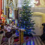 Muzycy w ludowych strojach grają w kościele obok choinki z kolorowymi światełkami. Tło zdobią złote i czerwone detale. Publiczność siedzi i robi zdjęcia.