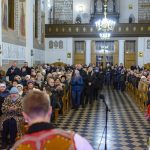 Ludzie zgromadzeni w kościele z jasnym wnętrzem, ozdobnymi freskami i żyrandolem. Na pierwszym planie muzycy w tradycyjnych strojach. Atmosfera jest radosna.
