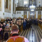 Grupa ludzi w kościele o jasnym wnętrzu, błyszczący żyrandol i muzycy w tradycyjnych strojach na pierwszym planie.