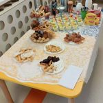 Na stole są talerze z ciastkami i filiżanki z deserami. W tle widać pudełka herbaty Lipton i dzbanek z kawą. Obrus jest biały z żółtą koronką.