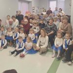 Grupa dzieci i dorosłych siedzi w sali, dzieci w niebieskich spódnicach i białych koszulach. W tle jasne ściany i okna. Kobieta trzyma dziecko, a inna robi zdjęcia telefonem.