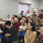 Grupa ludzi siedząca w sali z szarymi ścianami, jasnym sufitem. Uczestnicy ubrani są w różnorodne stroje. W tle widać tablice ogłoszeń.