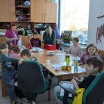 Messenger_creation_20657769742720861 Grupa dzieci przy stole, otoczona materiałami plastycznymi. Dwie dorosłe kobiety nadzorują pracę. Tło stanowią drewniane szafki i jasne okno.