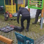 Dwóch mężczyzn pracuje przy placu zabaw z kolorowym domkiem. Obok narzędzia na kostce brukowej. W tle drzewa i szare niebo.