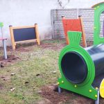 Zielono-fioletowy pociąg w piaskownicy, obok biała kuchenka. Tło z szarym murem i tablicami. Ziemia z zieloną trawą.