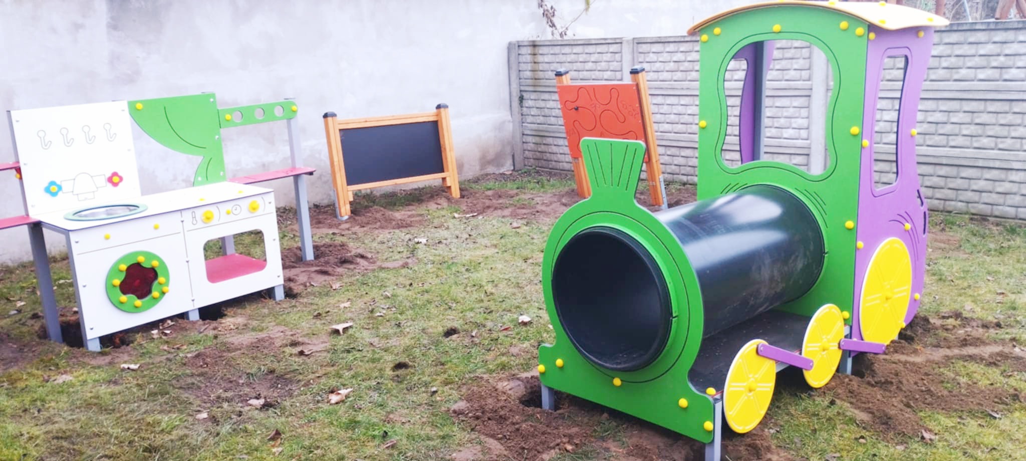Zielono-fioletowy pociąg w piaskownicy, obok biała kuchenka. Tło z szarym murem i tablicami. Ziemia z zieloną trawą.