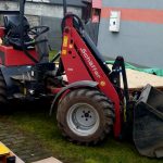 Na trawniku stoi czerwona ładowarka z dużą łyżką. W tle kolorowy plac zabaw i szare ogrodzenie. Na ziemi leży drewniana platforma z zielonymi elementami.