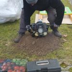 Mężczyzna w czapce obsługuje sprzęt na trawniku. Obok leży skrzynka z narzędziami. W tle widać ogrodzenie i drzewa.