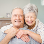 Na zdjęciu starsza para siedzi na kanapie, obejmując się i uśmiechając. Tło jest jasne, z drewnianą półką i książkami. Ubrania mają w odcieniach szarości i bieli.