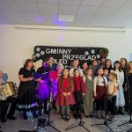 Grupa dzieci i dorosłych śpiewa kolędy na scenie; w tle czarne tło z napisem i dekoracje świąteczne. Tło jest jasne, a na podłodze są białe światełka. Po lewej stronie mężczyzna gra na akordeonie.