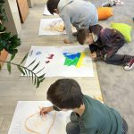 received_12223478098541061 Trzech chłopców rysuje na dużych kartkach papieru w jasnym pomieszczeniu z dywanem. Obok nich leżą kolorowe poduszki, a w tle widać regał z książkami. Paproć i drewniane panele dodają przytulności.