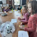 Grupa dzieci siedzi przy drewnianym stole, zajmując się projektami. Na stole leżą kolorowe arkusze i przedmioty, tło stanowią półki i okno. Dominują kolory naturalnego drewna, zieleni i różu.