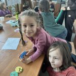 Grupa dzieci bawi się przy drewnianym stole. Dziewczynka w różowej bluzce trzyma kolorowy klocek. W tle widoczne są półki z książkami.