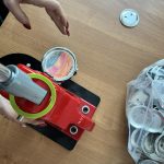 Na zdjęciu widoczna jest czerwona maszyna do robienia przypinek na drewnianym stole. Ręka z czerwonym lakierem na paznokciach wskazuje na kolorową przypinkę. Obok leży woreczek z metalowymi elementami.