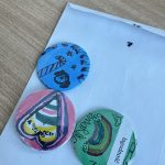 Trzy kolorowe przypinki leżą na białej kopercie. Niebieska z cukierkiem, różowa z sercem, zielona z tęczą. Tło drewniane.