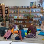 Grupa dzieci siedzi na dywanie w bibliotece, otoczona kolorowymi poduszkami. W tle są półki pełne książek. Jasne ściany i rośliny tworzą przytulną atmosferę.