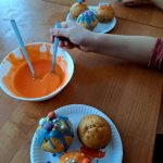 Trzy babeczki ozdobione kolorową polewą i posypkami leżą na talerzu. Obok miska z pomarańczowym lukrem. Dzieci dekorują ciastka przy drewnianym stole.