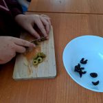 Osoba kroi zielone oliwki na desce do krojenia. Na drewnianym stole stoi biała miska z czarnymi oliwkami. Całość w jasnym świetle dziennym.