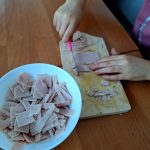 Dziecko kroi szynkę na desce, obok biała miska z plastrami. Tło to drewniany stół, różowa koszulka.