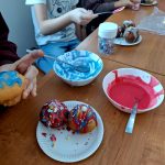 Na zdjęciu widać dzieci dekorujące babeczki kolorowym lukrem. Stół drewniany, miseczki z niebieskim i czerwonym lukrem. W tle siedzą dzieci w kolorowych ubraniach.