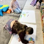 Dwie dziewczynki rysują kredkami na dużych kartkach. Tło jest jasne, z regałem z książkami i poduszkami na dywanie. Obok widać plecaki i fotele.