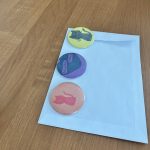 Na drewnianym stole leży biała koperta z trzema kolorowymi przypinkami. Przypinki mają rysunki i kolory: żółty z kotem, fioletowy oraz różowy z kotem. Tło jest jednolite, drewniane.